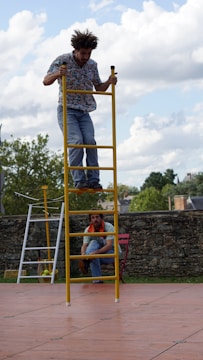 A person is balancing on top of a yellow ladder, positioned vertically above another person who is crouching towards the bottom. The background features a natural setting with trees and a stone wall, under a partly cloudy sky.