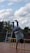 A person is performing a handstand on a yellow ladder placed on a wooden platform. They are wearing blue jeans and flippers while balancing upside down. The background shows a cloudy sky and greenery, with a stone wall and a ladder nearby.