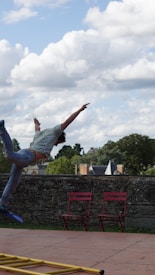 A person mid-air during a jump, against a backdrop of a cloudy sky. Red folding chairs are positioned against a stone wall, and a yellow object is visible at the bottom of the image. The scene takes place outdoors, surrounded by greenery and distant rooftops.