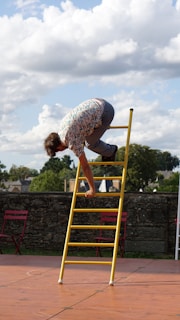 A person performing a maneuver on a yellow ladder, positioned horizontally in an outdoor setting. The individual is crouched on the second rung from the top wearing a patterned shirt and gray pants. The backdrop contains a stone wall, trees, and a partly cloudy sky.