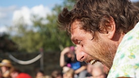 A person with a beard is captured mid-laughter in an outdoor setting with a blurred background of people and trees. The individual is wearing a shirt with a light green and yellow pattern, and the natural light enhances their expression.