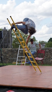 A person is performing an acrobatic jump over a yellow ladder set up on a wooden stage. Another person sits on a red chair in the background, set against a stone wall and an outdoor setting with trees and clouds.
