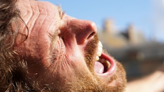 A close-up of a person's face with expressions indicating intense emotion, possibly yelling or expressing strong feelings. The lighting is bright and the person's features, including a beard and open mouth, are prominently visible.
