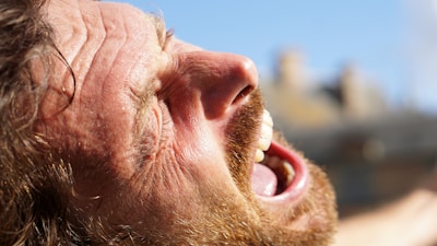 A close-up of a person's face with expressions indicating intense emotion, possibly yelling or expressing strong feelings. The lighting is bright and the person's features, including a beard and open mouth, are prominently visible.