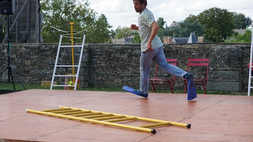 A person wearing flippers on their feet appears to be playfully skipping or balancing on a wooden platform. A yellow ladder is laid on the platform. In the background, there is a stone wall with trees and a clear blue sky. Red folding chairs and other miscellaneous objects are visible, suggesting an outdoor setting.