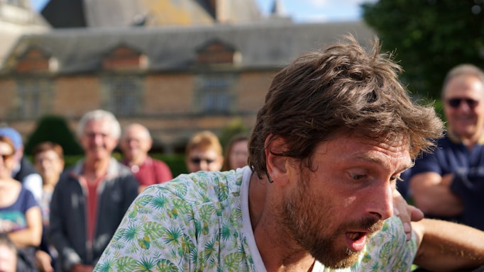 A man with messy hair and a surprised expression stands in the foreground, wearing a shirt with a green leafy pattern. Behind him, a group of people is gathered, some wearing sunglasses, in an outdoor setting with a large building in the background.