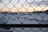 A chain-link fence is visible in the foreground with a blurred snowy landscape and a marina with docks in the background. The sun is setting, casting a warm glow across the sky and creating a contrast with the cold environment.