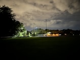 A nighttime scene with a sports field illuminated by light poles and a small building emitting warm light. The sky is partly cloudy, casting a dim, diffused glow. Dense trees border the left side of the field, creating a dark contrast against the lighter elements.