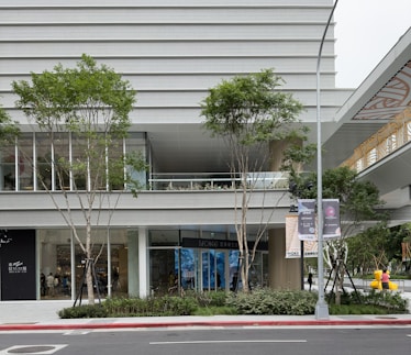 A modern commercial building exterior featuring large glass windows and vertical architectural lines. The street in front of the building is bordered by young trees and neatly trimmed shrubs. On the right, there are two large, colorful sculptures resembling bears, adding a playful element to the scene. A wide sidewalk and a streetlamp with banners contribute to the urban setting, while a covered walkway with decorative designs is visible above.
