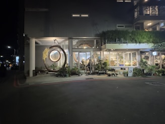 A storefront at night with large glass windows displaying a variety of plants inside. A unique circular swing made of wood with a translucent center is positioned at the entrance. The building facade is modern with plenty of greenery and lighting, creating a warm and inviting atmosphere.