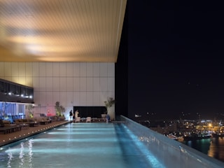 An illuminated rooftop infinity pool at night, adjacent to a modern building with a wooden ceiling. The city lights twinkle in the background, creating a serene atmosphere. People are seen on the deck, some walking and others relaxing on lounge chairs, while potted plants add a touch of greenery.