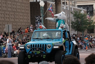 A vibrant parade scene with a jeep decorated in blue driving down a street. A person in a dolphin costume is standing in the vehicle's back, engaging with the crowd. Numerous spectators, some dressed in themed jerseys, are seated alongside the street, watching the parade. American and Ohio flags are visible among the surrounding urban architecture.