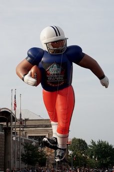 A giant inflatable balloon of an American football player is displayed in a parade setting. The figure is depicted wearing a football uniform with a helmet and holding a football. Flags and buildings are visible in the background, along with a clear sky.