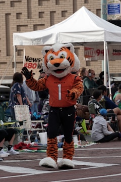 A person in a tiger mascot costume with orange and black stripes and wearing a jersey that says 'BENGALS' with the number 1 is performing in front of a seated crowd. The setting appears to be an outdoor community or sports event with a white tent and people sitting on chairs and blankets in the background.