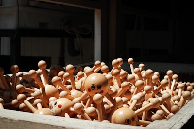 A collection of wooden toys with spherical heads and multiple stick-like arms, each adorned with a smiling face. The toys appear to be in a bin, crowded together, with sunlight casting shadows on them.