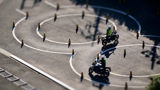 Motorcyclists navigate through a series of winding paths marked by traffic cones on a spacious asphalt surface. The riders appear to be in a training or practice setting, wearing helmets and safety vests. The sunlight casts crisp shadows on the ground, highlighting the serpentine pattern of the chalk lines and cones.