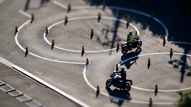 Motorcyclists navigate through a series of winding paths marked by traffic cones on a spacious asphalt surface. The riders appear to be in a training or practice setting, wearing helmets and safety vests. The sunlight casts crisp shadows on the ground, highlighting the serpentine pattern of the chalk lines and cones.