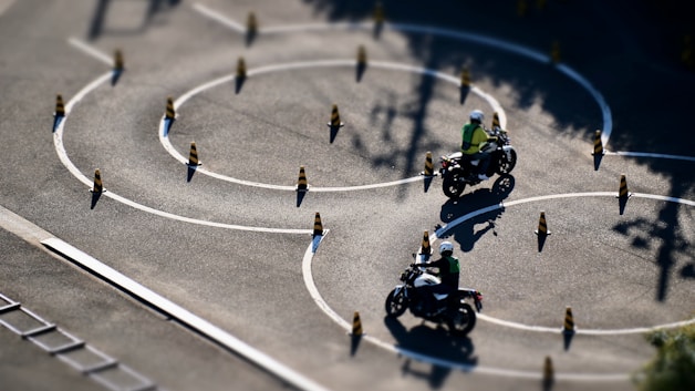 Motorcyclists navigate through a series of winding paths marked by traffic cones on a spacious asphalt surface. The riders appear to be in a training or practice setting, wearing helmets and safety vests. The sunlight casts crisp shadows on the ground, highlighting the serpentine pattern of the chalk lines and cones.