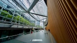 A vast, modern architectural space with a high glass ceiling and expansive windows. The right side features a large, curved, textured wall, while the left side displays lush greenery visible through glass panels. People are seen walking on the gray floor, providing a sense of scale.