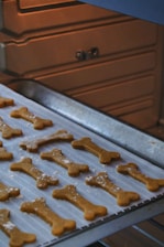 Baking tray lined with parchment paper holds several bone-shaped cookies inside an oven. The cookies appear golden-brown and lightly dusted with flour.
