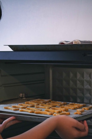 A person is placing a baking tray with letter-shaped cookies into an oven. The surrounding area has dim lighting, and a towel is resting on the edge of the tray.