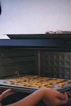 A person is placing a baking tray with letter-shaped cookies into an oven. The surrounding area has dim lighting, and a towel is resting on the edge of the tray.