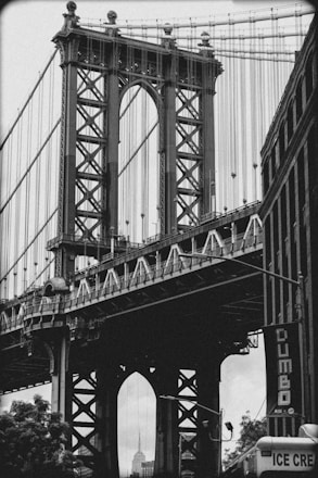 A black and white photograph of the Manhattan Bridge with its intricate ironwork and suspension cables prominently featured. The Empire State Building is visible through the bridge's supports. The photograph captures the urban architecture and street level atmosphere of the Dumbo neighborhood.