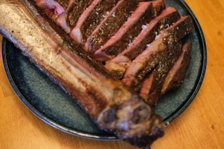 Sliced grilled meat with a charred crust is arranged on a blue plate. The meat appears juicy with a pink inner color and is sprinkled with coarse salt. The plate is placed on a wooden surface.