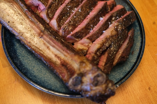 Sliced grilled meat with a charred crust is arranged on a blue plate. The meat appears juicy with a pink inner color and is sprinkled with coarse salt. The plate is placed on a wooden surface.