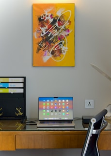 A modern workspace with a laptop on a wooden desk, displaying a screen full of app icons. Above the desk hangs a vibrant abstract painting with yellow, orange, and various colors, featuring dynamic white brush strokes. To the left of the laptop is a partition with phrases like 'TO KNOW' and 'TO DO'. A black office chair is partially visible, and a desk lamp is on the right side.