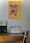 A modern workspace with a laptop on a wooden desk, displaying a screen full of app icons. Above the desk hangs a vibrant abstract painting with yellow, orange, and various colors, featuring dynamic white brush strokes. To the left of the laptop is a partition with phrases like 'TO KNOW' and 'TO DO'. A black office chair is partially visible, and a desk lamp is on the right side.