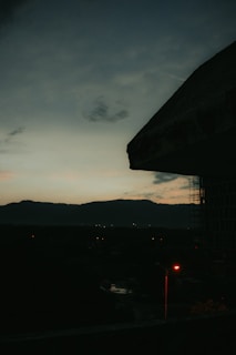 A dark, moody cityscape at dusk with a barely visible outline of mountains in the distance. The sky transitions from a deep blue to muted orange near the horizon. A prominent silhouette of a building occupies the right side of the frame, with a dimly glowing red streetlamp in the foreground.