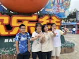 A group of four young people stands in front of a large colorful sign featuring both Chinese and English text, celebrating a 25th anniversary. They are smiling and making peace signs with their hands. The background includes flowers, festive decorations, and a vibrant amusement park atmosphere.