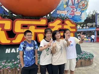 A group of four young people stands in front of a large colorful sign featuring both Chinese and English text, celebrating a 25th anniversary. They are smiling and making peace signs with their hands. The background includes flowers, festive decorations, and a vibrant amusement park atmosphere.