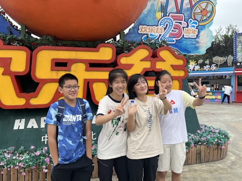 A group of four young people stands in front of a large colorful sign featuring both Chinese and English text, celebrating a 25th anniversary. They are smiling and making peace signs with their hands. The background includes flowers, festive decorations, and a vibrant amusement park atmosphere.
