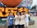 A group of four young people stands in front of a large colorful sign featuring both Chinese and English text, celebrating a 25th anniversary. They are smiling and making peace signs with their hands. The background includes flowers, festive decorations, and a vibrant amusement park atmosphere.