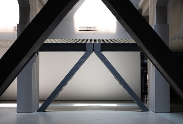 A geometric, architectural interior with large, angular steel beams forming a triangular shape. The beams are set against a plain, light-colored wall and are part of a larger industrial structure, possibly inside a building or a factory. The lighting is subdued, creating soft shadows and highlights.