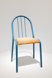 A simple chair with a blue metal frame and a seat made of light-colored woven material. The chair has a rounded back with vertical metal bars.
