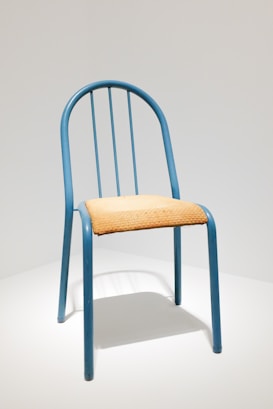 A simple chair with a blue metal frame and a seat made of light-colored woven material. The chair has a rounded back with vertical metal bars.