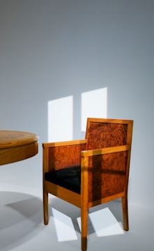 A wooden chair with a dark cushioned seat and intricately patterned backrest sits next to a wooden table. The wall in the background is illuminated by soft, diffused sunlight casting rectangular patterns on the surface.