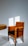 A wooden chair with a dark cushioned seat and intricately patterned backrest sits next to a wooden table. The wall in the background is illuminated by soft, diffused sunlight casting rectangular patterns on the surface.