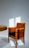 A wooden chair with a dark cushioned seat and intricately patterned backrest sits next to a wooden table. The wall in the background is illuminated by soft, diffused sunlight casting rectangular patterns on the surface.