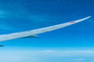 A large airplane wing extending across the frame with a logo on it, set against a clear blue sky with scattered clouds below.
