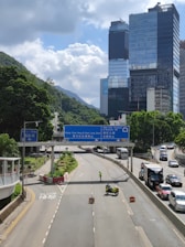 A multi-lane road with a motorcycle and a person blocking one section, likely for road maintenance or control. Several cars are driving on the open lanes, and large buildings with reflective glass facades are seen in the background against a backdrop of lush green hills. Traffic signs in both English and Chinese are present, indicating directions.