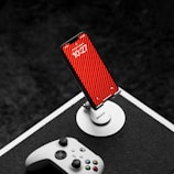A smartphone on a stand with a red striped wallpaper is placed on a table. In the foreground, an Xbox controller lies nearby, suggesting a gaming setup. The background is dark, which provides contrast to the bright colors of the phone and the controller.
