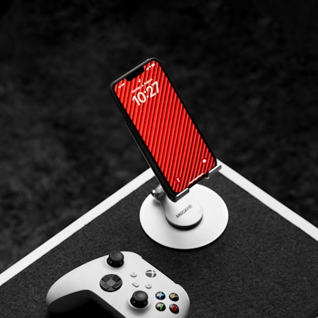 A smartphone on a stand with a red striped wallpaper is placed on a table. In the foreground, an Xbox controller lies nearby, suggesting a gaming setup. The background is dark, which provides contrast to the bright colors of the phone and the controller.