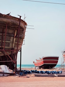 Two large boats are situated on a sandy shore near an ocean. The boat in the foreground is unfinished, revealing its wooden structure and scaffolding. The second boat in the background is painted red and white, resting on supports. Several blue barrels and construction materials are scattered around, indicating a boat-building or repair site.