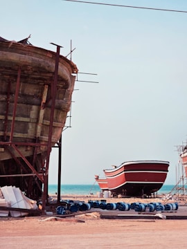 Two large boats are situated on a sandy shore near an ocean. The boat in the foreground is unfinished, revealing its wooden structure and scaffolding. The second boat in the background is painted red and white, resting on supports. Several blue barrels and construction materials are scattered around, indicating a boat-building or repair site.