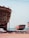 Two large boats are situated on a sandy shore near an ocean. The boat in the foreground is unfinished, revealing its wooden structure and scaffolding. The second boat in the background is painted red and white, resting on supports. Several blue barrels and construction materials are scattered around, indicating a boat-building or repair site.
