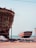 Two large boats are situated on a sandy shore near an ocean. The boat in the foreground is unfinished, revealing its wooden structure and scaffolding. The second boat in the background is painted red and white, resting on supports. Several blue barrels and construction materials are scattered around, indicating a boat-building or repair site.